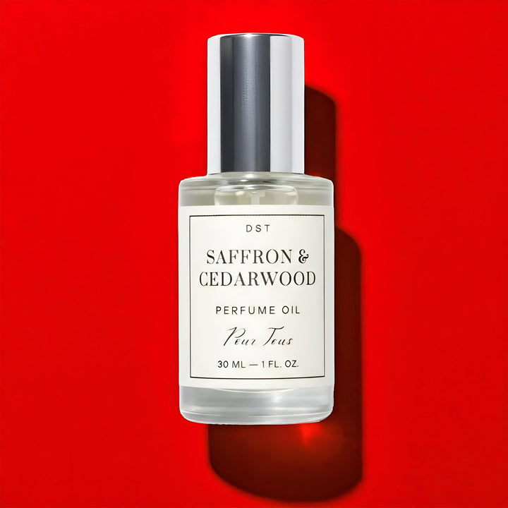 DST Fragrances Perfume oil bottle labeled 'Saffron & Cedarwood' laying on a light wood surface with a red ribbon in the background