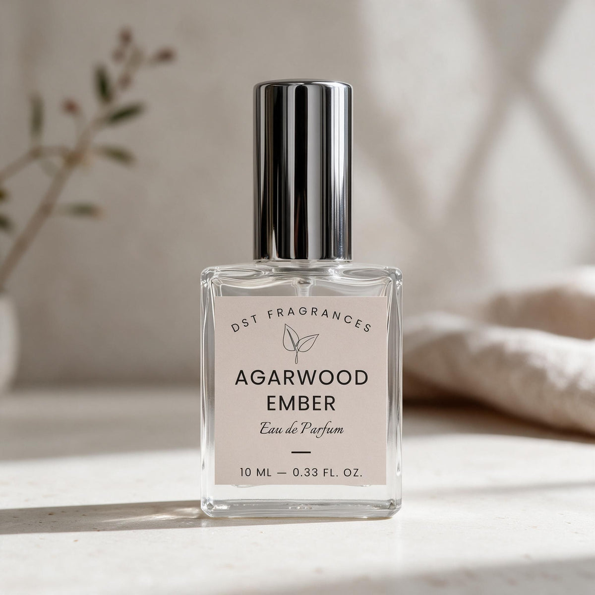 A 10 mL bottle of DST Fragrances Agarwood Ember Eau de Parfum, featuring a clear glass bottle, a silver metallic cap, and a beige label, set on a light marble surface with a soft-focus background.