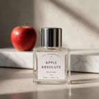 A clear glass bottle of dsT Fragrances 'Apple Absolute' Eau de Toilette with a silver cap and white label, set on a marble surface next to a red apple.