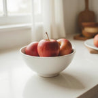 Three red apples in a white ceramic bowl on a light countertop near a window, with a softly blurred kitchen background.