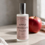 A bottle of Apple Absolute Body Splash by dst Fragrances, featuring a frosted glass bottle with a silver cap and a light pink label, set on a stone surface next to a red apple.