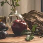Red apple, plums, a piece of large weathered wood and greenery sitting on a wood tabletop.