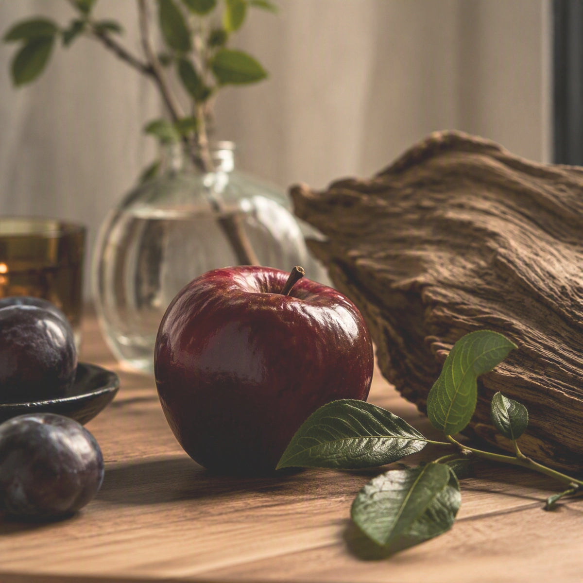 Red apple, plums, a piece of large weathered wood and greenery sitting on a wood tabletop.