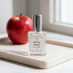 A 10 mL bottle of DST FRAGRANCES Apple Noir Eau de Toilette sits on a white marble tray next to a fresh red apple, with a soft-focus window background.