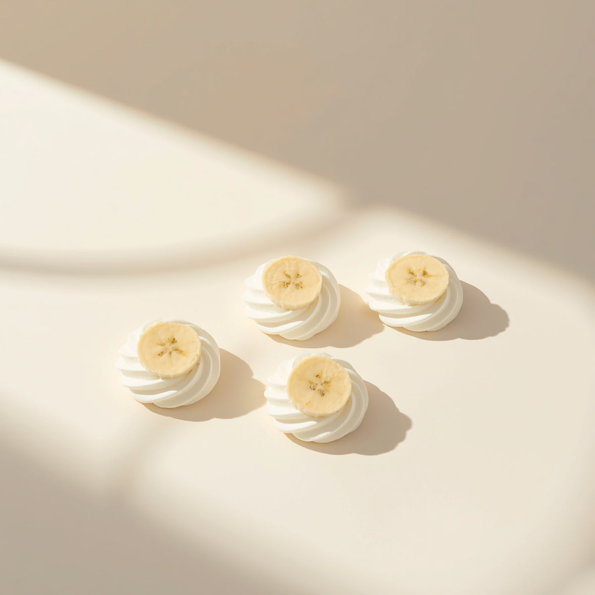 Four small white whipped cream or frosting dollops, each topped with a thin sliced banana, arranged on a warm cream surface with soft diagonal sunlight and shadows.