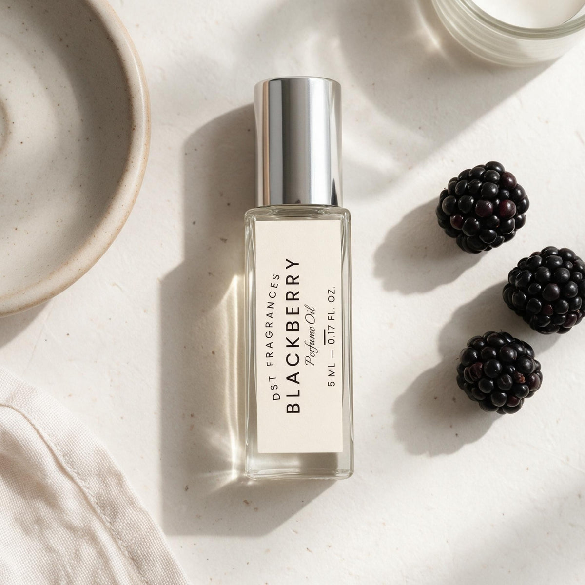 5ml bottle of Blackberry scented perfume oil by DST Fragrances against an ivory surface surrounded by blackberries and neutral colored linens and pottery.
