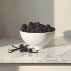 A white ceramic bowl filled with fresh blackberries on a white marble countertop, with vanilla beans placed beside it in soft natural light.