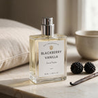 A clear rectangular perfume bottle labeled 'Blackberry Vanilla' by Dst Fragrances, set on a marble surface with blackberries, a vanilla bean, and soft home decor in the background.