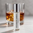 A 10 mL bottle of Bourbon Ember Perfume Oil by DST Fragrances stands in the foreground on a light marble surface, with a blurred glass of amber whiskey in the background.
