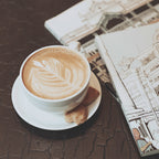 A white ceramic cup of cappuccino with leaf latte art sits on a matching saucer, with two cookies beside it and illustrated books or magazines on a dark textured tabletop.