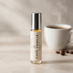 A small glass roll-on bottle labeled 'Café Vanille' by DST Fragrances stands on a light surface with a blurred white coffee cup and coffee beans in the background.