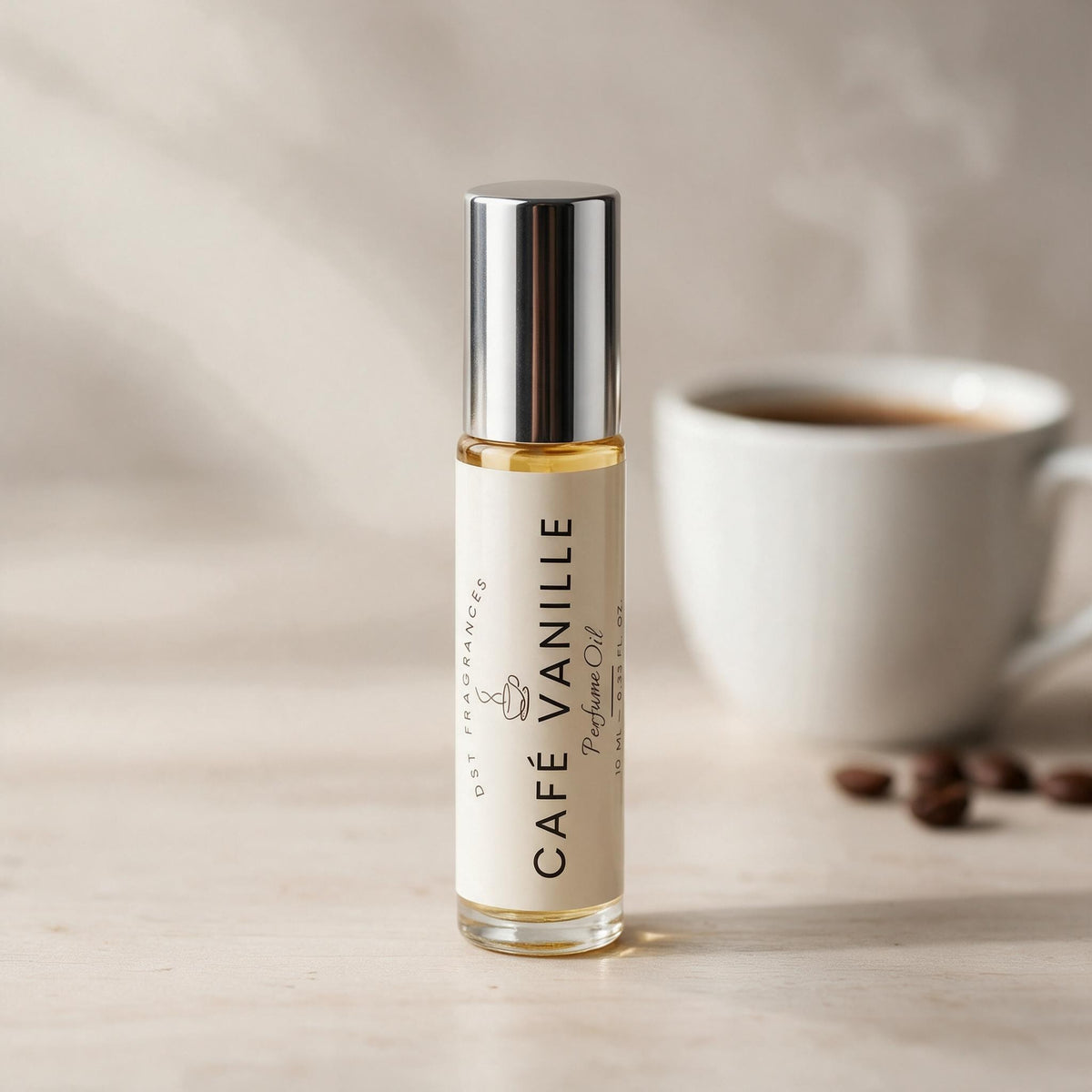 A small glass roll-on bottle labeled 'Café Vanille' by DST Fragrances stands on a light surface with a blurred white coffee cup and coffee beans in the background.