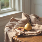 A beige fringed throw blanket draped over a wooden table next to a window, with a whole and halved fig resting on top.