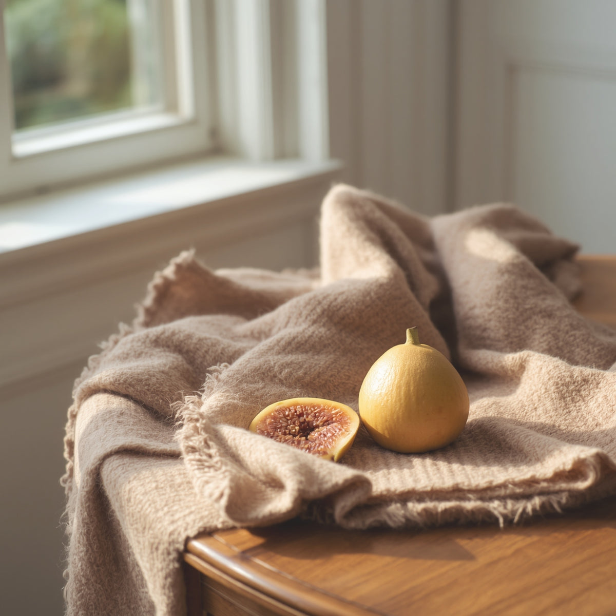 A beige fringed throw blanket draped over a wooden table next to a window, with a whole and halved fig resting on top.