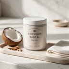 A jar of Coconut Santal body butter by Dst Fragrances sits on a marble tray next to a halved coconut, cinnamon sticks, and a folded cream towel. The scene is styled with warm, natural light and a soft-focus background.