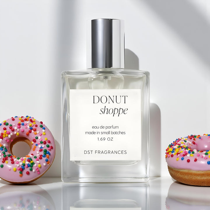 50ml bottle of Donut Shoppe Eau de Parfum by dst fragrances sitting on a glossy white surface with 2 donuts covered in pink frosting and rainbow sprinkles against a light grey background.