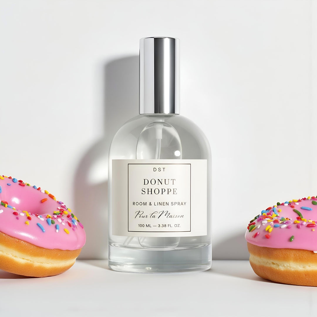 100ml bottle of Donut Shoppe Room Linen Spray by DST Fragrances sitting on a white surface with 2 donuts covered in bright pink frosting and rainbow sprinkles against a light grey backdrop.