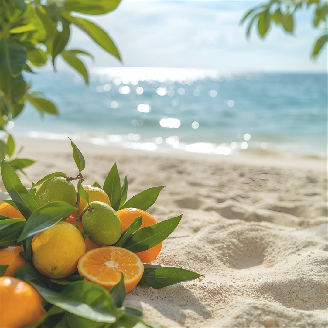 A cluster of fresh oranges, limes, and green leaves arranged on sandy beach ground with the ocean and sky blurred in the background.