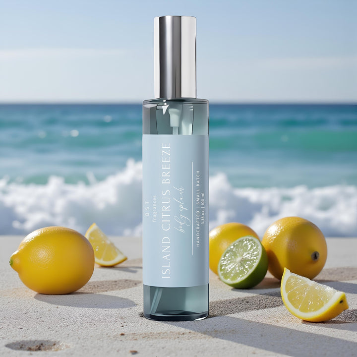 100ml bottle of Island citrus breeze body splash by DST Fragrances sitting on a sandy beach next to lemons and limes with the ocean and blue sky in the background.