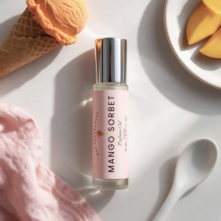 A clear glass bottle of Mango Sorbet fragrance with a pale pink label and reflective silver cap, styled on a white surface with a waffle cone, peach ice cream scoop, pink cloth, white spoon, and peach slices.