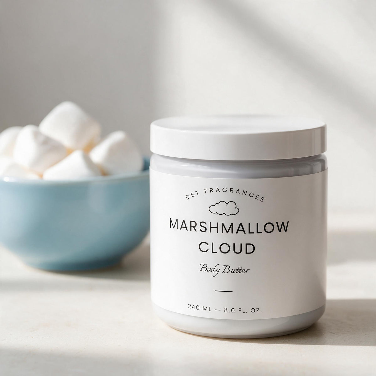 A white jar of DST Fragrances Marshmallow Cloud Body Butter sits on a light surface with a blue bowl of marshmallows blurred in the background.