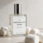 A clear glass perfume bottle labeled 'Marshmallow Cream' by Dst Fragrances, with a reflective silver cap and a white label, styled with marshmallows on a light beige surface.