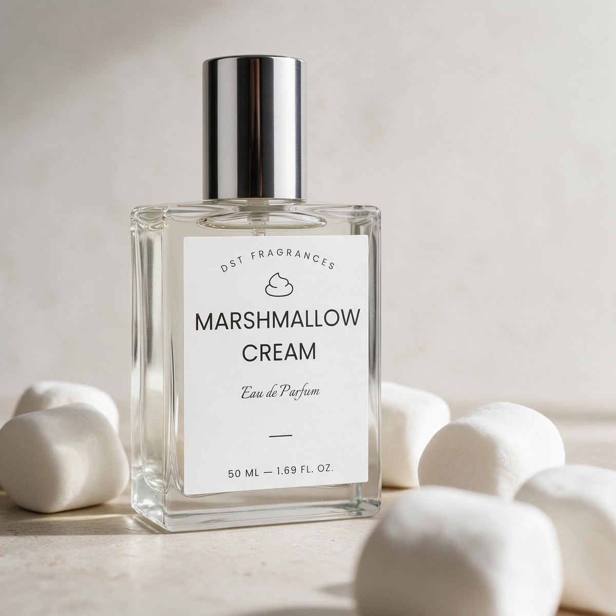 A clear glass perfume bottle labeled 'Marshmallow Cream' by Dst Fragrances, with a reflective silver cap and a white label, styled with marshmallows on a light beige surface.