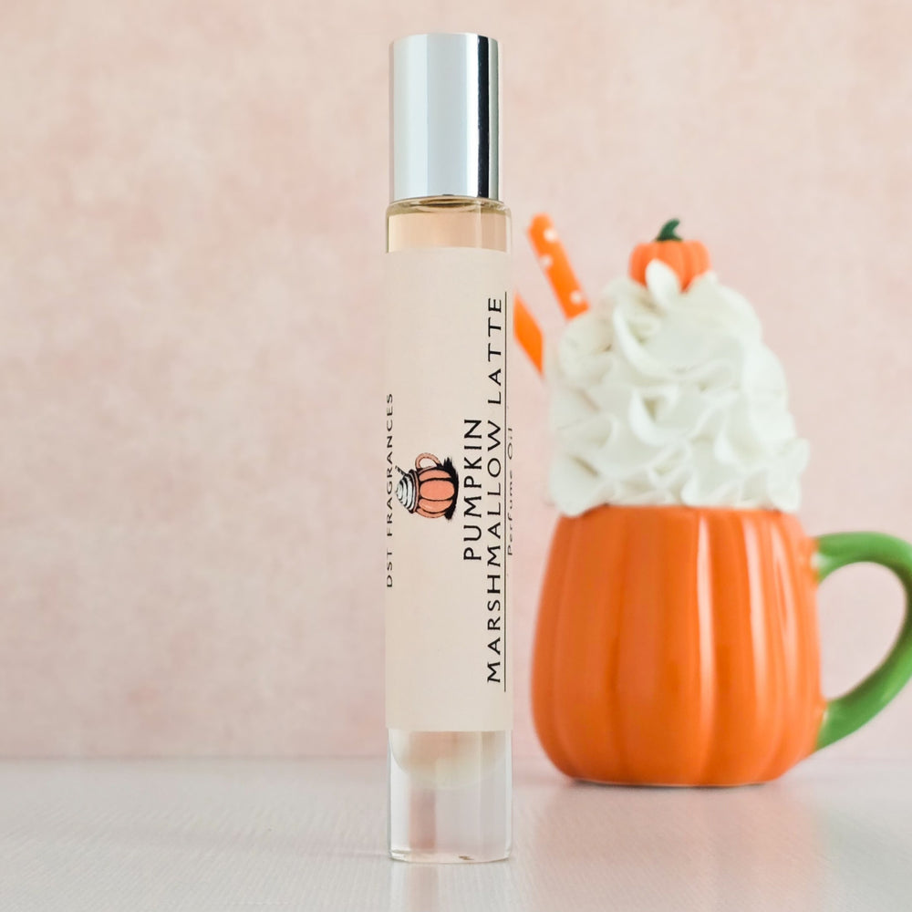 Perfume bottle labeled 'Pumpkin Marshmallow Latte' next to a pumpkin-shaped mug with whipped cream and a candy corn on a light pink background.