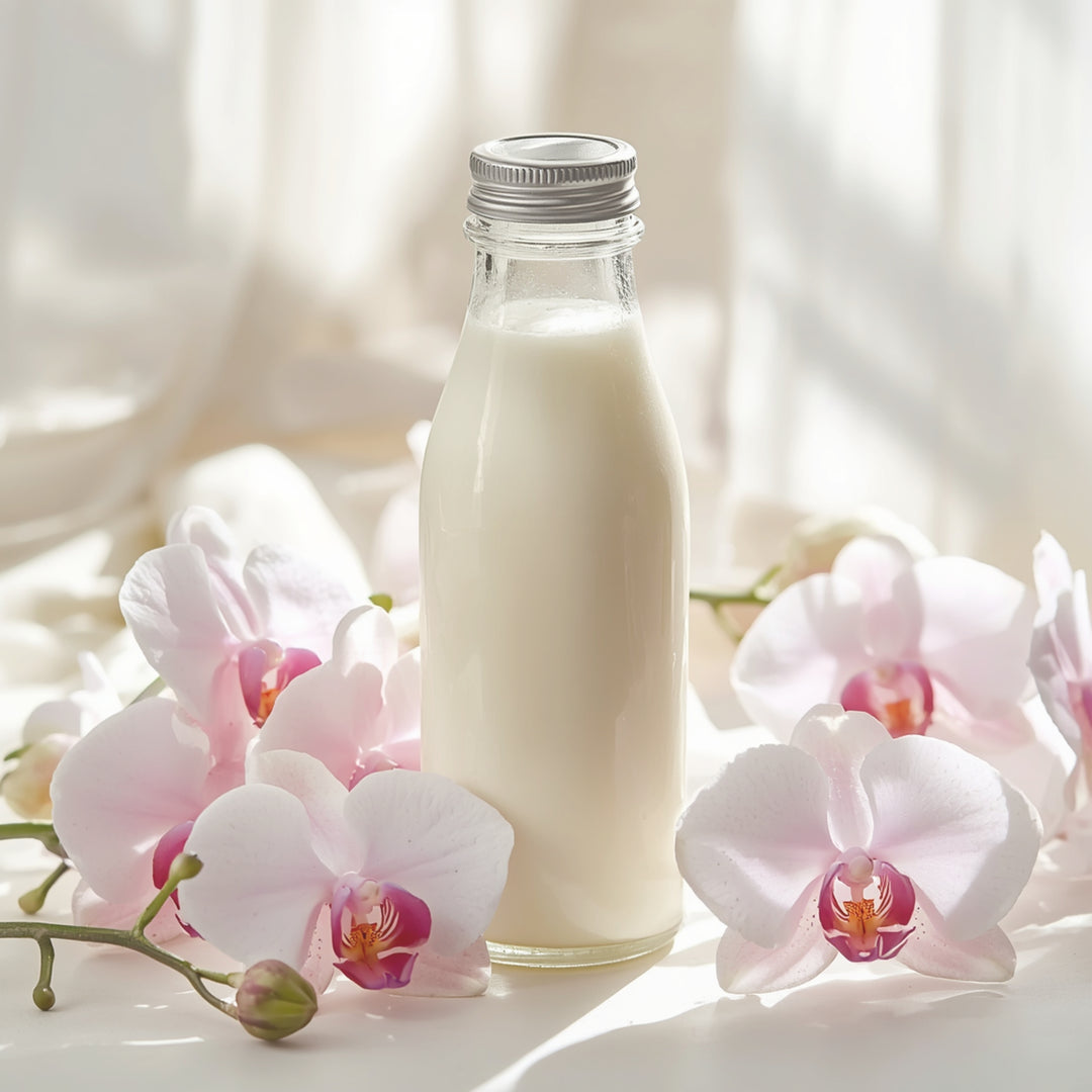 A clear glass bottle filled with white milk or milk substitute, standing upright and surrounded by pale pink orchid flowers on a softly lit white surface with a warm neutral background.