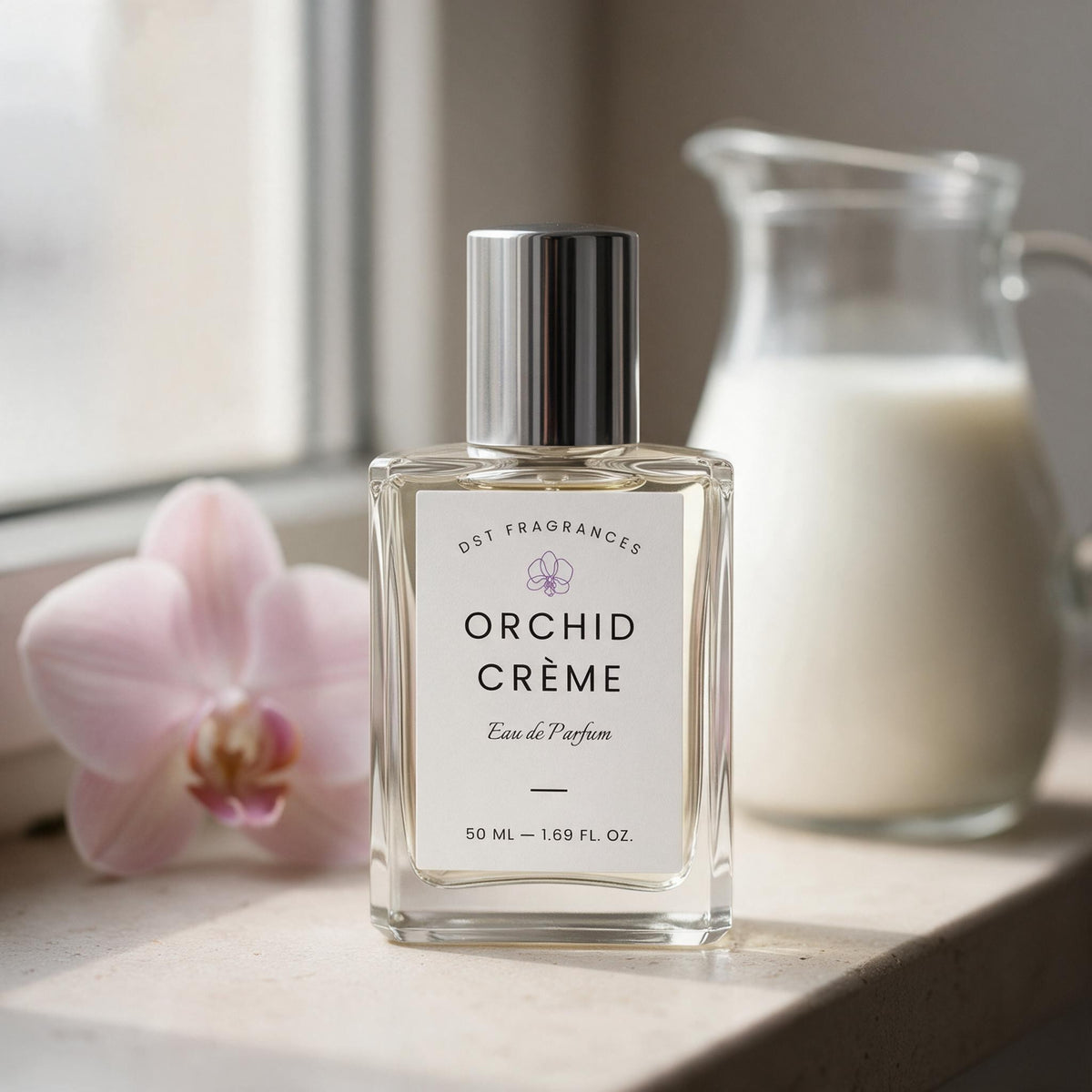 A clear glass perfume bottle labeled 'ORCHID CRÈME' by dst Fragrances, with a silver cap and white label, set on a windowsill with a pink orchid flower and a glass pitcher in the background.