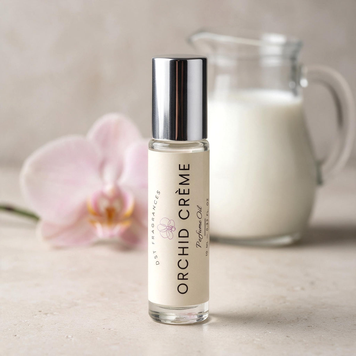 A clear glass perfume oil bottle labeled 'ORCHID CRÈME' with a silver cap stands on a light stone surface. A pale pink orchid flower and a blurred glass pitcher of milk appear in the background.