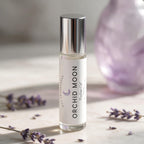 A clear glass bottle of orchid oil with a reflective silver cap and a white label reading 'ORCHID MOON' and 'Ostremacenced', standing on a light marble surface with scattered lavender sprigs and a soft purple blurred background object.