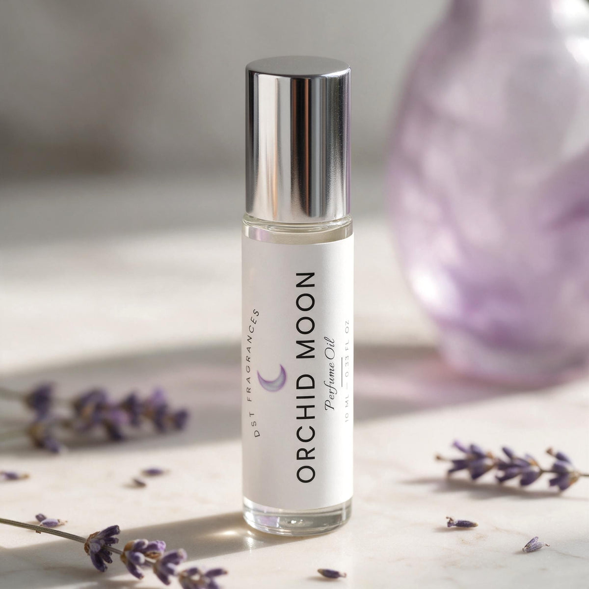 A clear glass bottle of orchid oil with a reflective silver cap and a white label reading 'ORCHID MOON' and 'Ostremacenced', standing on a light marble surface with scattered lavender sprigs and a soft purple blurred background object.