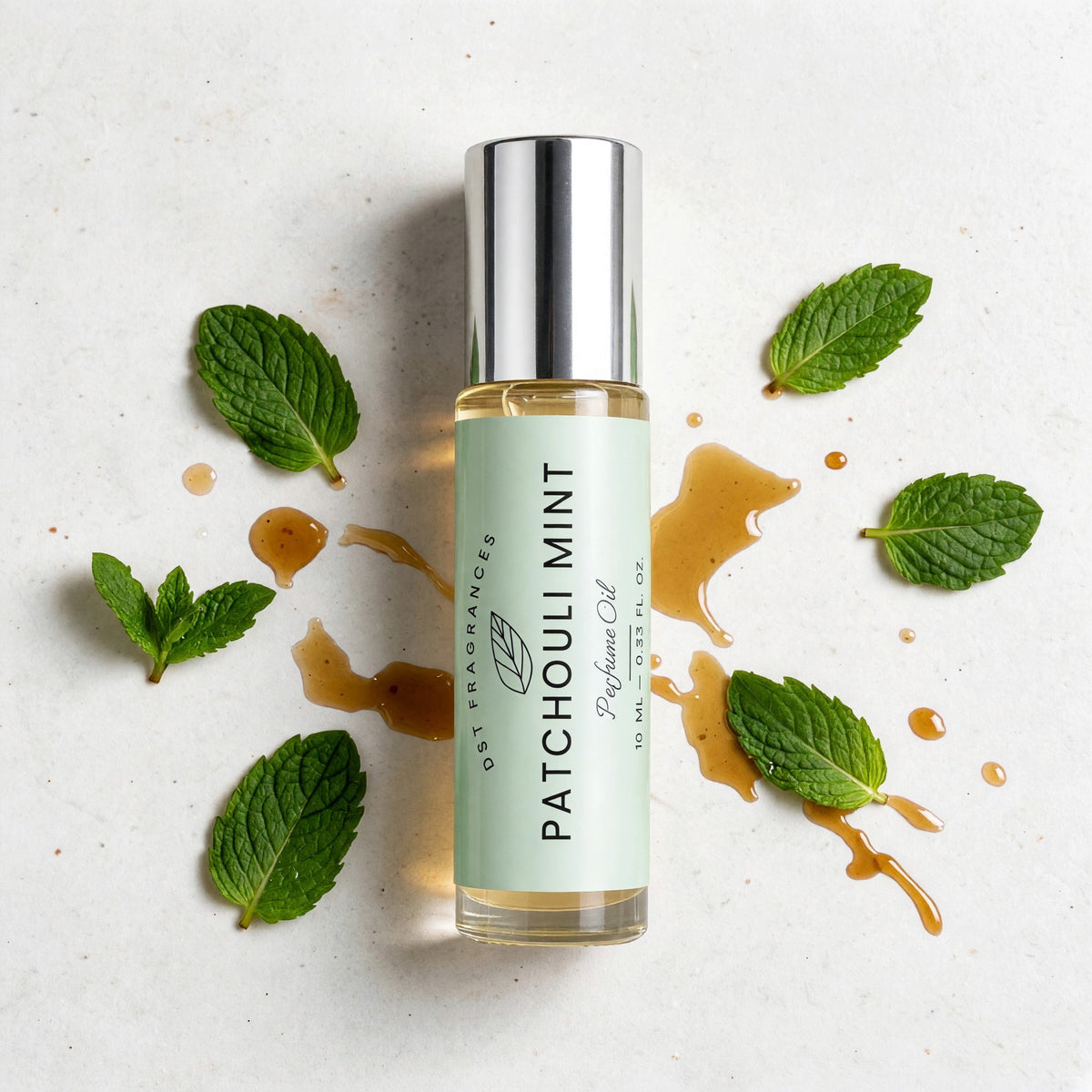 A clear glass perfume oil bottle with a silver cap and a pale mint-green label reading 'Patchouli Mint' and 'Dst Fragrances,' styled on a light surface with mint leaves and amber syrup around it.