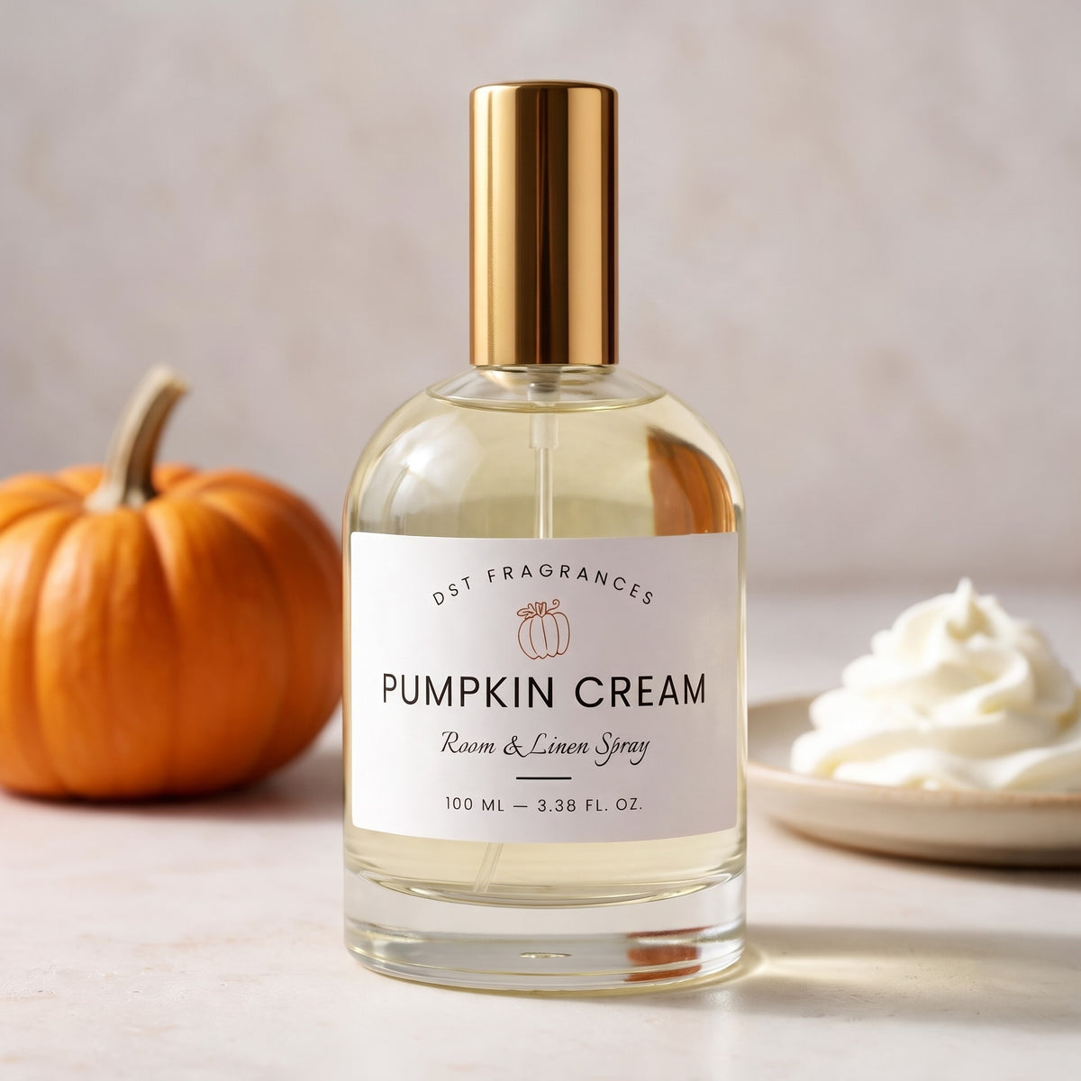 A bottle of DST Fragrances Pumpkin Cream Room & Linen Spray stands on a light surface, with a small orange pumpkin on the left and a dollop of whipped cream on a plate on the right. The bottle has a clear glass body, pale yellow liquid, a white label, and a metallic gold cap.