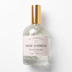 A 100 ml glass bottle of Dst Fragrances Sage Cypress Room & Linen Spray, featuring a clear glass body, a pale pink label, and a metallic gold atomizer cap, set against a clean white background.