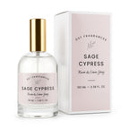 A clear glass bottle of DST Fragrances Sage Cypress Room & Linen Spray with a gold cap, shown next to its white and pale pink packaging box.
