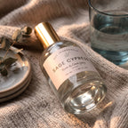 A bottle of Sage Cypress room and linen spray by DST Fragrances, featuring a clear glass body, a gold cap, and a cream label, styled on a beige knit blanket next to a glass of water and a small dish with eucalyptus.