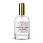 A clear glass bottle of Santo Cedar Room & Linen Spray by Dst Fragrances, featuring a white label and a shiny gold spray nozzle, set against a plain white background.
