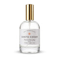 A clear glass bottle of Santo Cedar Room & Linen Spray by Dst Fragrances, featuring a white label and a shiny gold spray nozzle, set against a plain white background.
