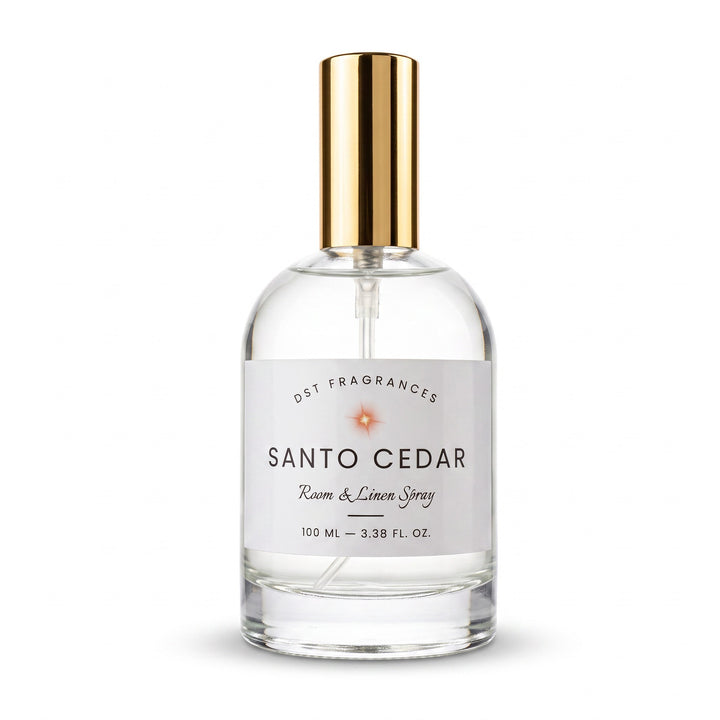 A clear glass bottle of Santo Cedar Room &amp; Linen Spray by Dst Fragrances, featuring a white label and a shiny gold spray nozzle, set against a plain white background.