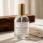 A clear glass bottle of Santo Cedar Room & Linen Spray by DST Fragrances, featuring a white label and a shiny gold cap, set on a linen surface with a wooden block and a folded cream towel in the background.