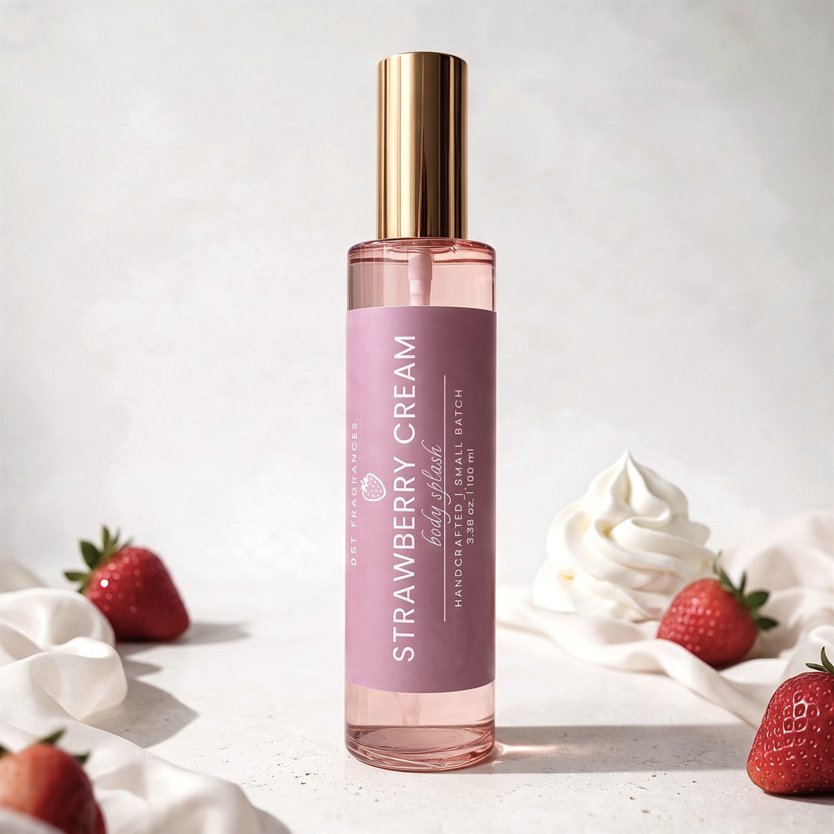 A bottle of Strawberry Cream body spray by DST Fragrances, featuring a dusty pink label and a metallic gold cap, styled on a light gray surface with strawberries and whipped cream.