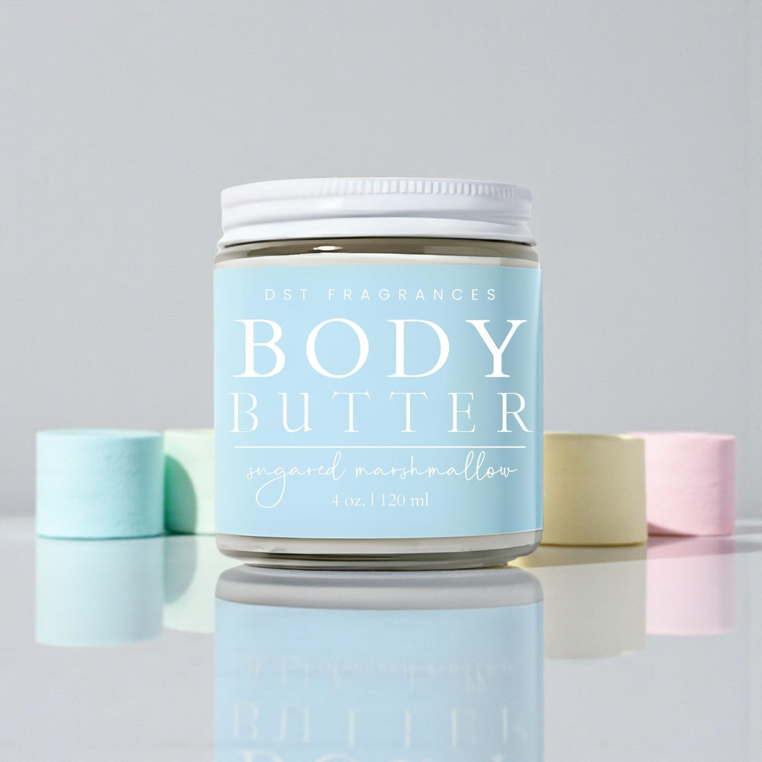 120ml jar of Sugared Marshmallow Scented body butter by DST Fragrances sitting on a reflective white surface surrounded by pastel colored marshmallows against a pale grey background.