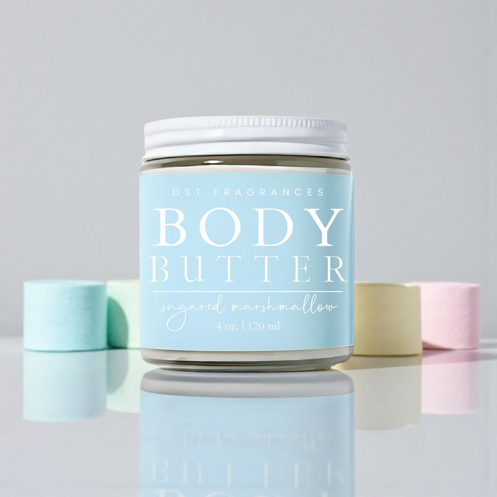 120ml jar of Sugared Marshmallow Scented body butter by DST Fragrances sitting on a reflective white surface surrounded by pastel colored marshmallows against a pale grey background.