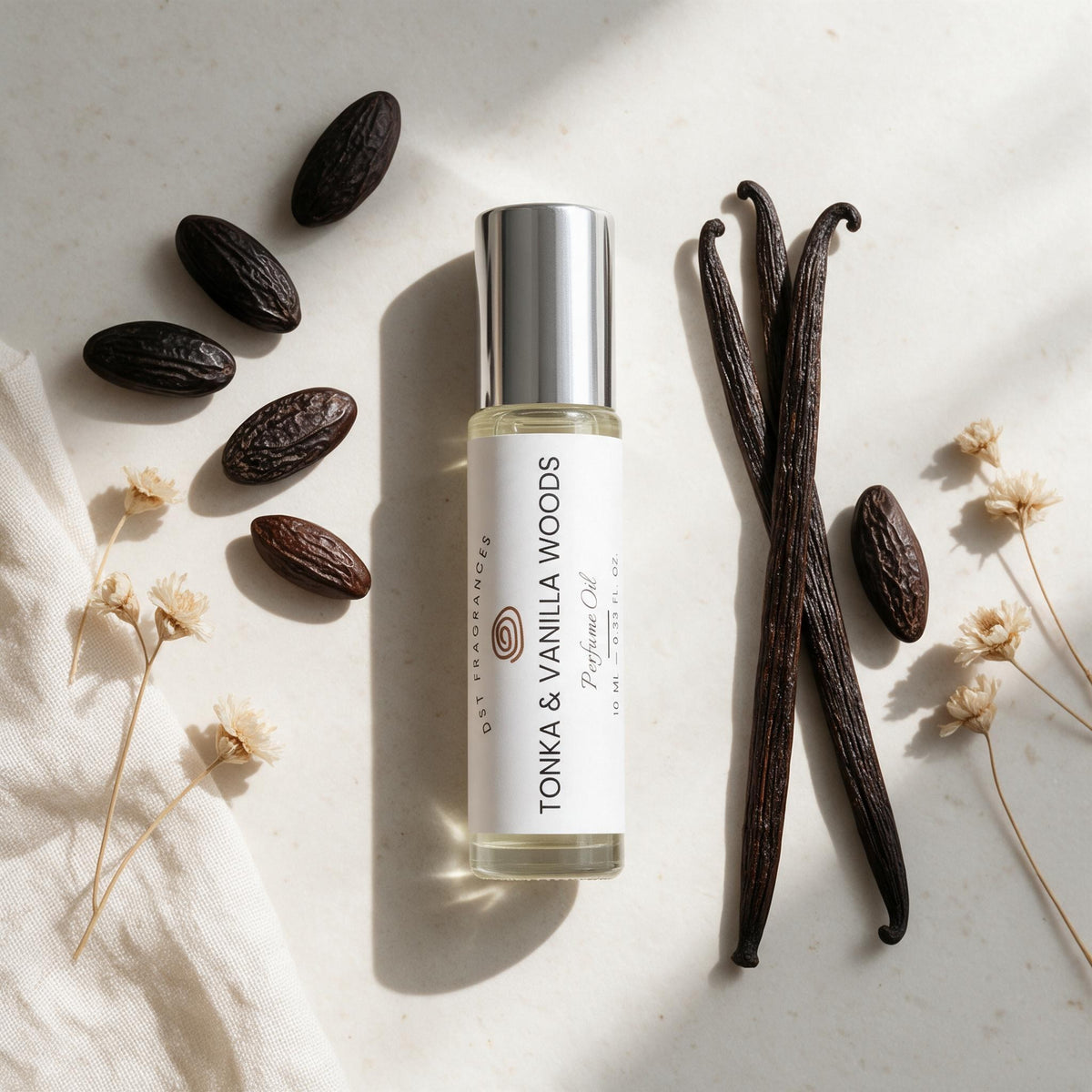 A clear cylindrical bottle of oil with a silver cap and a white label reading 'TONKA & VANILLA WOODS' is arranged on a light stone surface with vanilla beans, cocoa beans, and dried flowers. This is a styled flat-lay product shot with no person or real environment, so subject_type is Product/Packaging.