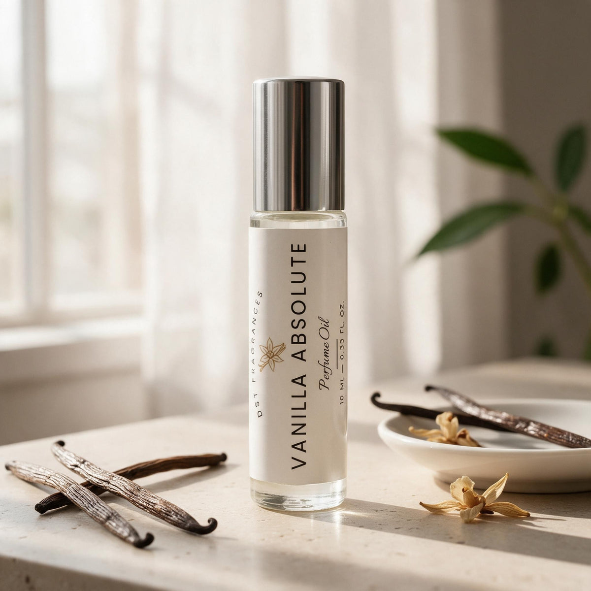 A clear cylindrical perfume oil bottle labeled 'VANILLA ABSOLUTE' with a silver cap, standing on a light stone surface near vanilla beans and a small dish, with a softly blurred indoor background.