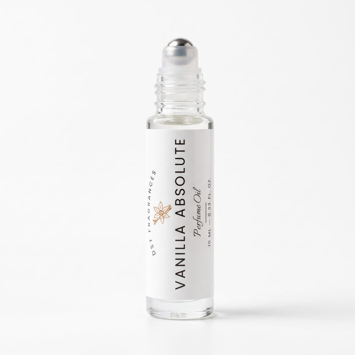 A 10 mL clear glass rollerball bottle of Vanilla Absolute perfume oil by DST Fragrances, featuring a white label with black text and a small gold starburst logo, topped with a silver cap, set against a clean white background.