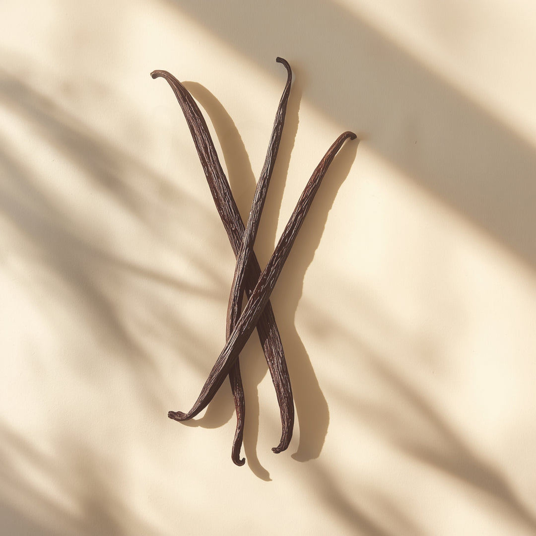 Three dark brown vanilla beans arranged in an X shape on a warm beige surface with soft leaf shadows.