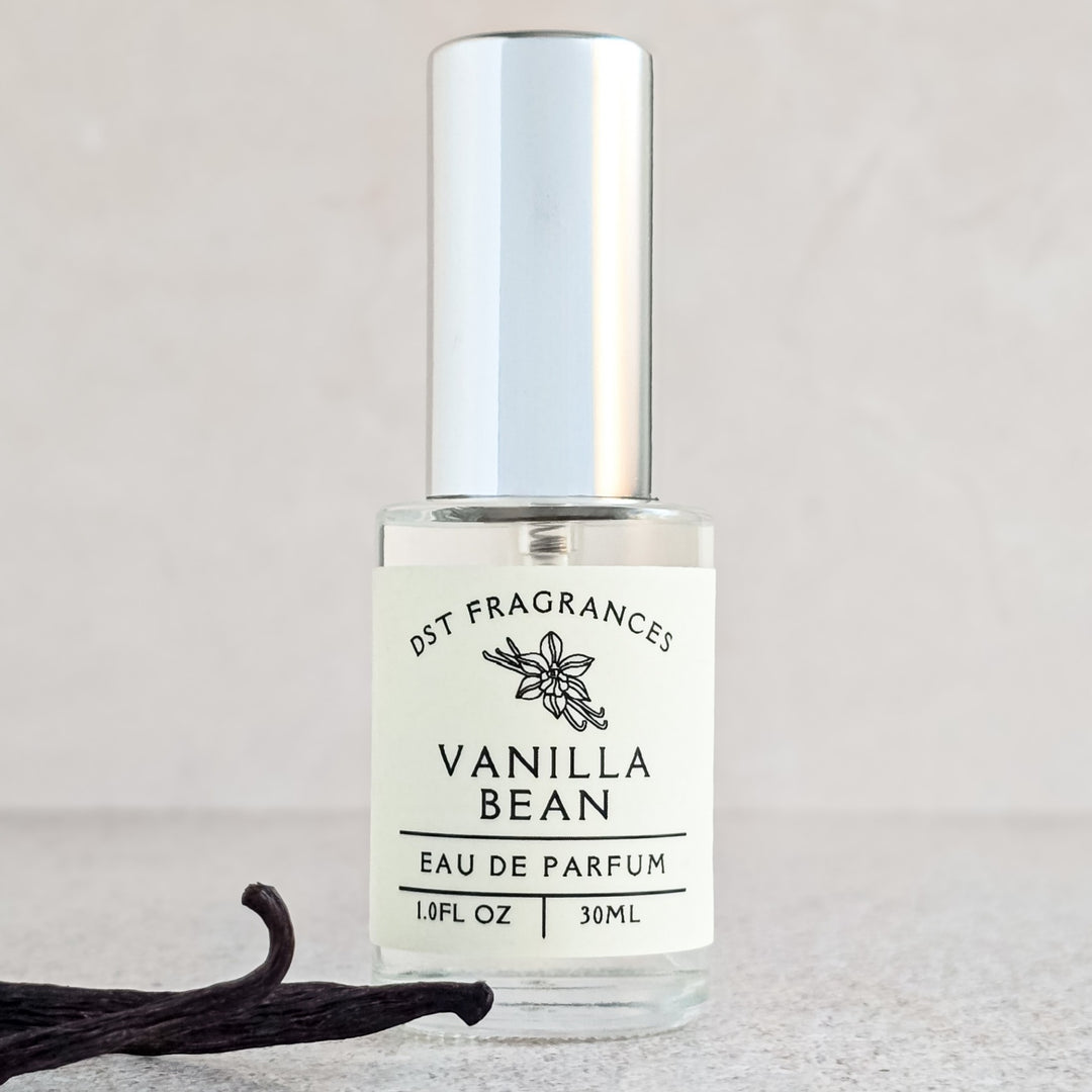Vanilla Bean eau de parfum bottle on a decorative stand with a vanilla bean next to it against a beige background.